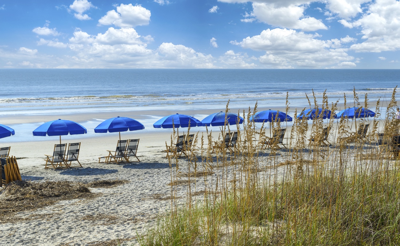 Pristine beach with blue umbrellas and lounge chairs awaiting guests along the sandy shoreline under a beautiful cloudy sky.