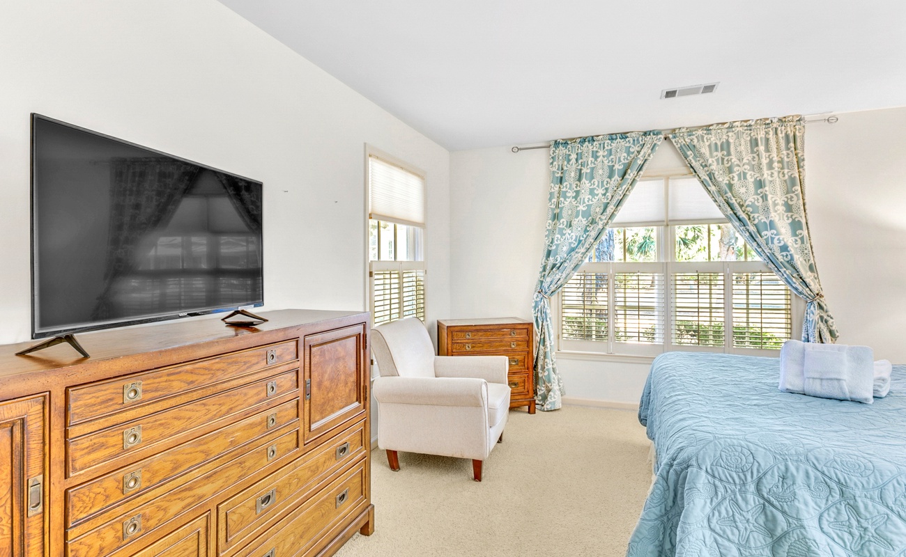 Unwind in your spacious bedroom with cozy blue bedding, large TV for relaxing evenings, and charming curtains framing peaceful garden views.