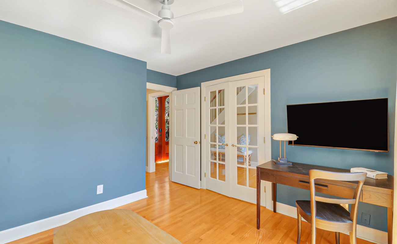 Your private workspace features a sleek desk, comfortable chair, and wall-mounted TV, all bathed in natural light from elegant French doors.