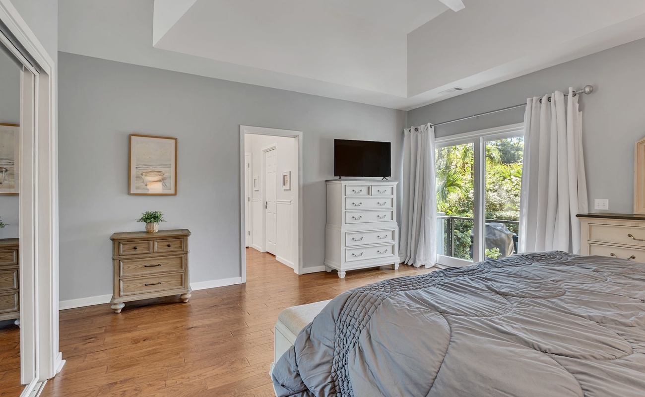 Relax in your spacious bedroom featuring elegant gray walls, warm hardwood floors, and large windows overlooking lush greenery.