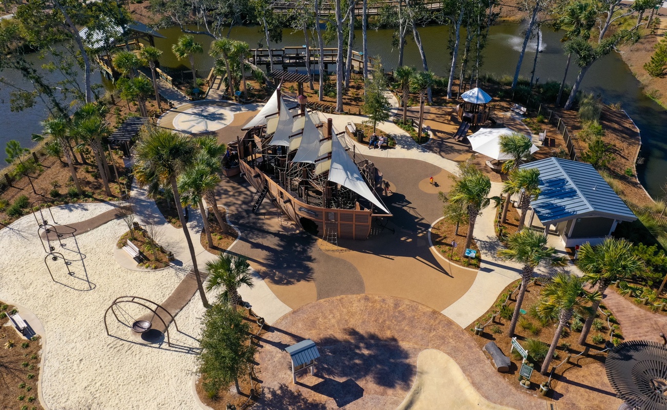 Aerial view of an elaborate adventure playground featuring a pirate ship centerpiece, climbing structures, and tropical landscaping near the waterfront.