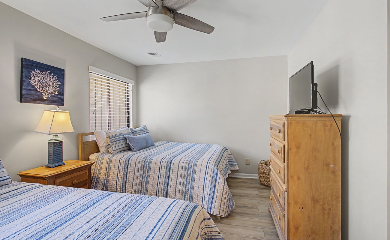 Relax in your peaceful twin bedroom with coastal decor, gentle ceiling fan, and warm natural light streaming through windows.