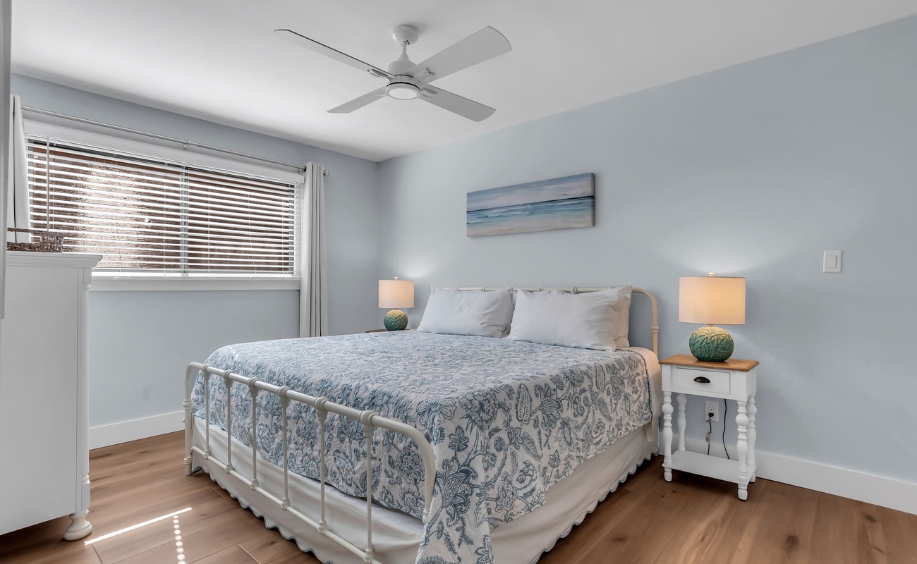 A welcoming guest bedroom designed with soft coastal tones and calming natural light. The comfortable bed is dressed in airy blue-and-white linens, complemented by matching nightstands and warm bedside lighting.