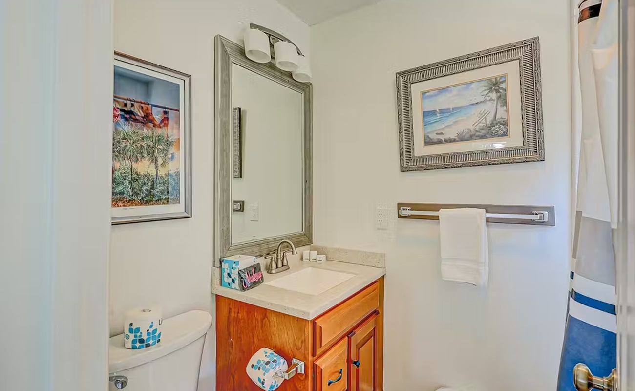 Your private bathroom features warm wood vanity, coastal artwork, and fresh towels—everything you need to refresh after beach adventures.