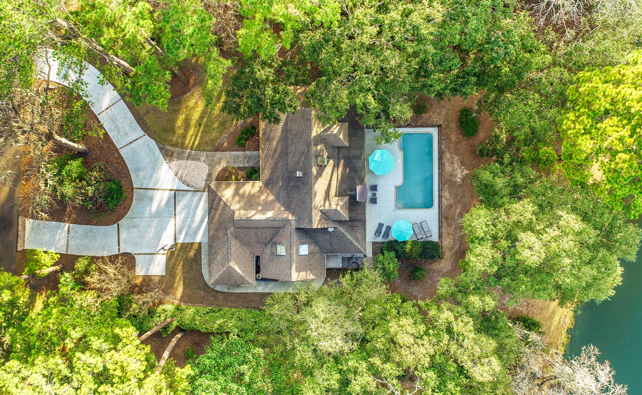 Aerial view of a tranquil lakeside property featuring a swimming pool, spacious outdoor areas, and winding driveway surrounded by lush greenery.