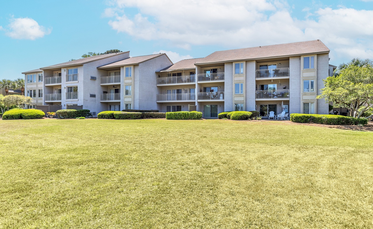 Multi-story vacation rental complex with private balconies overlooking expansive green lawn areas under bright blue skies.