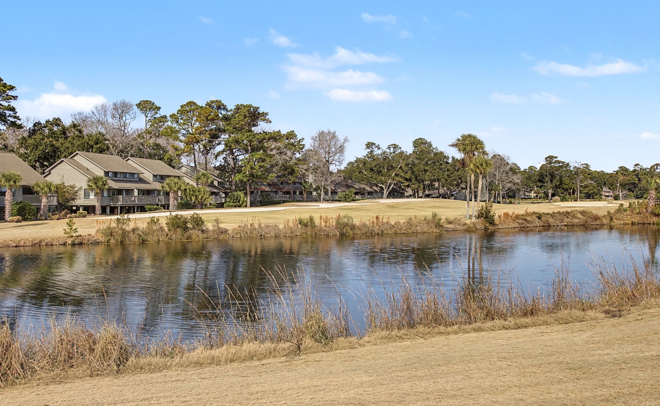 Golf course community featuring pristine fairways alongside a tranquil water hazard, surrounded by mature trees and residential properties.