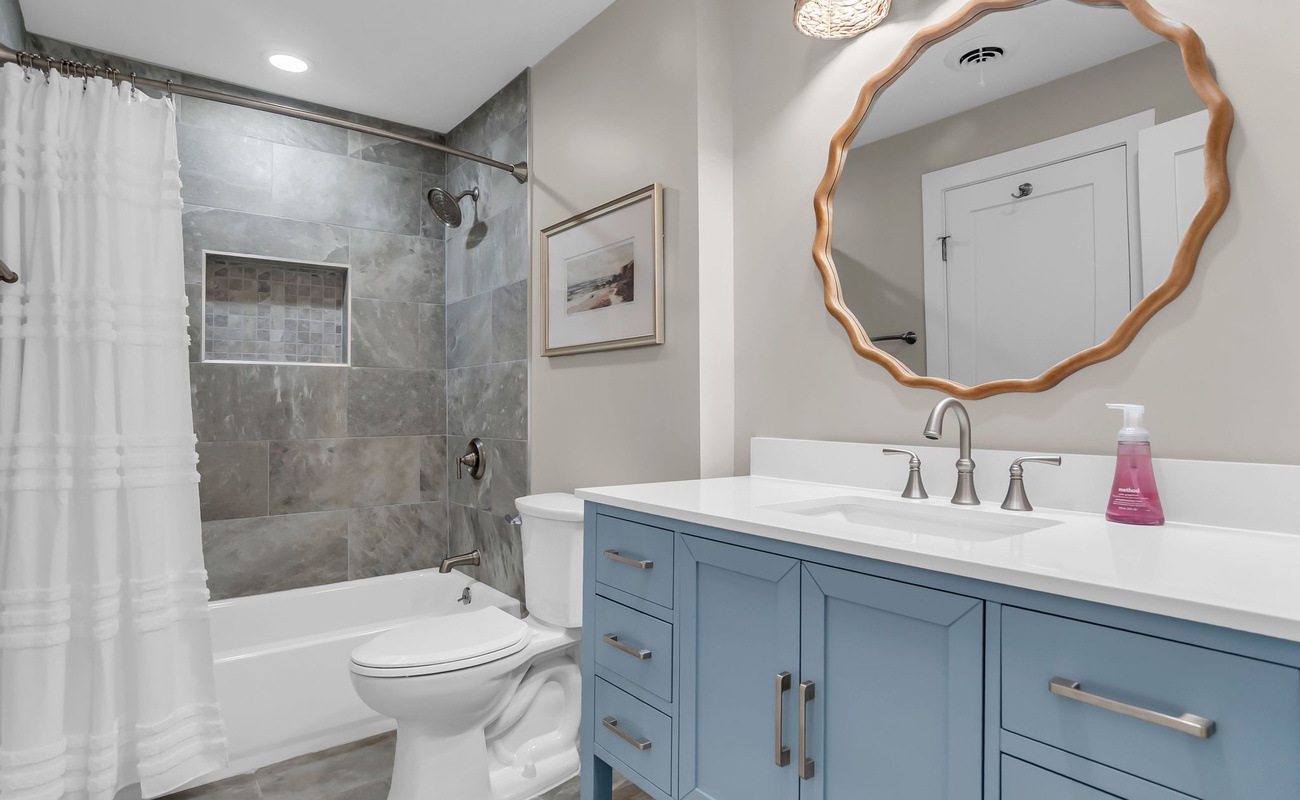 A beautifully updated bathroom featuring a custom blue vanity with polished hardware and a striking round mirror that adds a touch of coastal elegance. The tiled tub and shower combination showcases modern finishes and a built-in niche for convenience.
