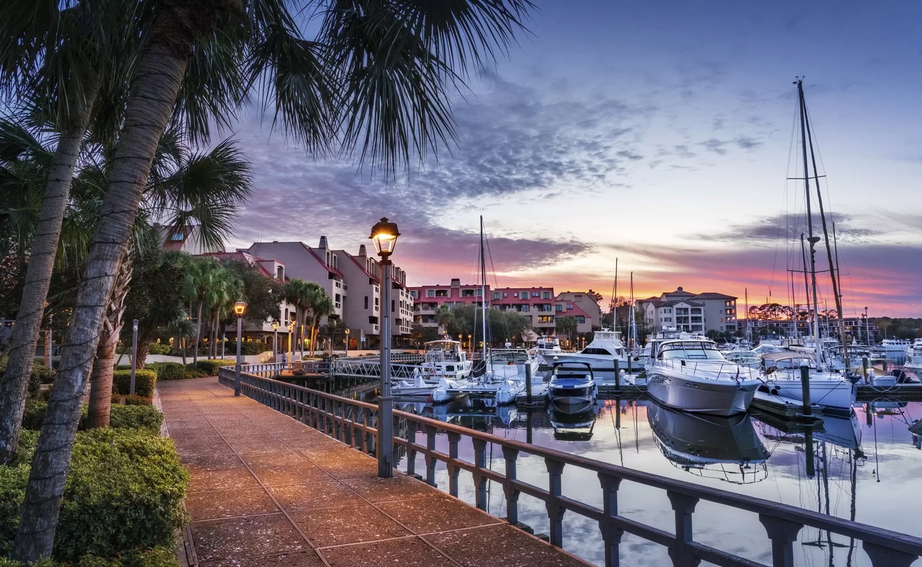 Waterfront marina at twilight with boats docked along the harbor, palm trees, and colorful buildings creating a picturesque coastal setting.