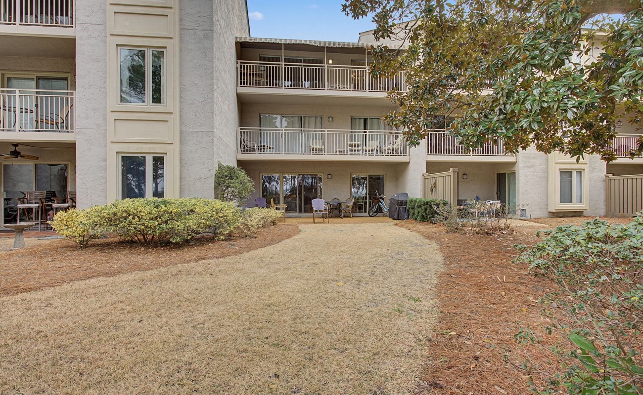 Multi-story condominium complex with ground-floor patios and balconies, surrounded by mature landscaping and open lawn areas.