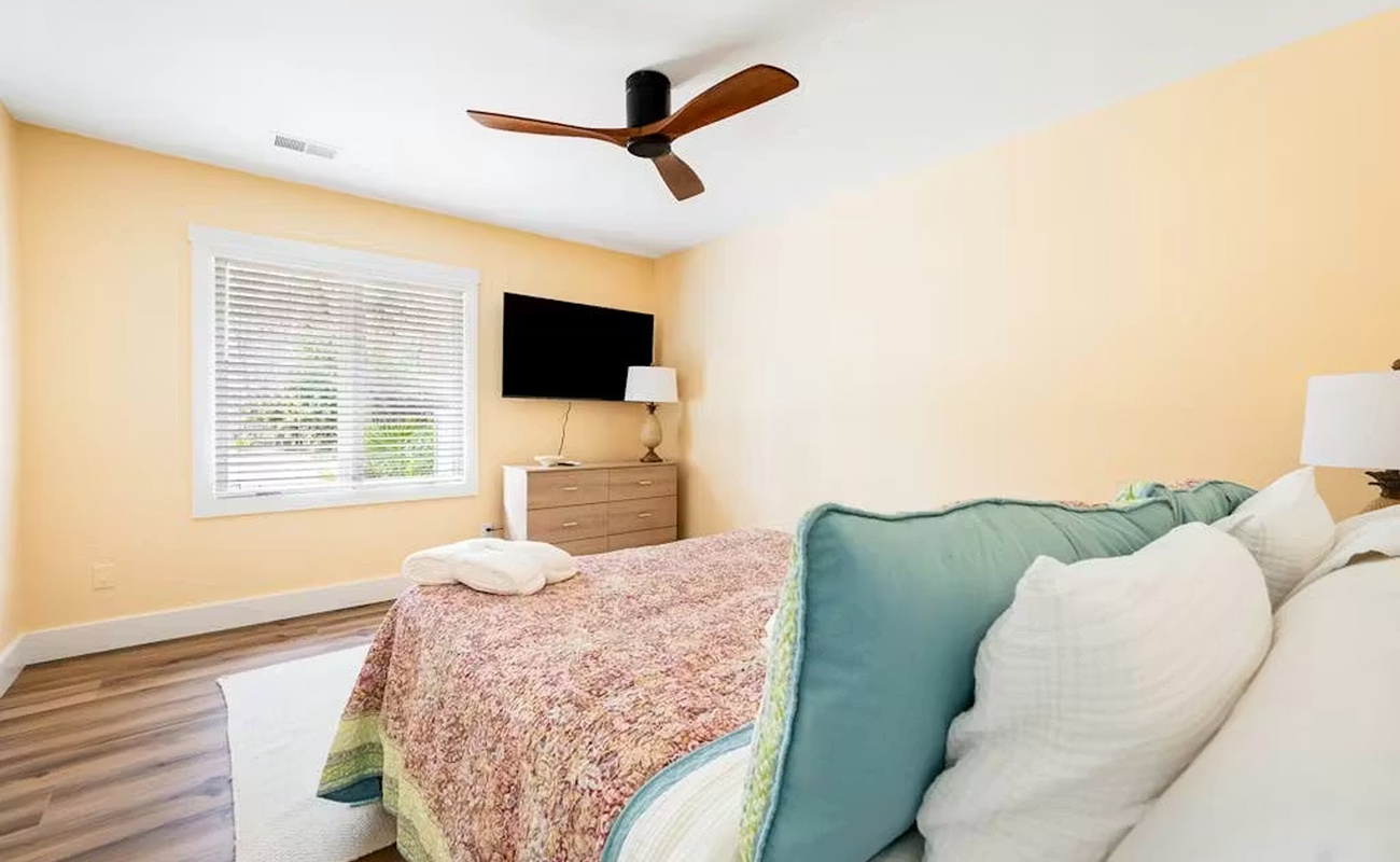Rest peacefully in your bright, airy bedroom featuring cozy linens, ceiling fan comfort, and tranquil garden views through blinds.