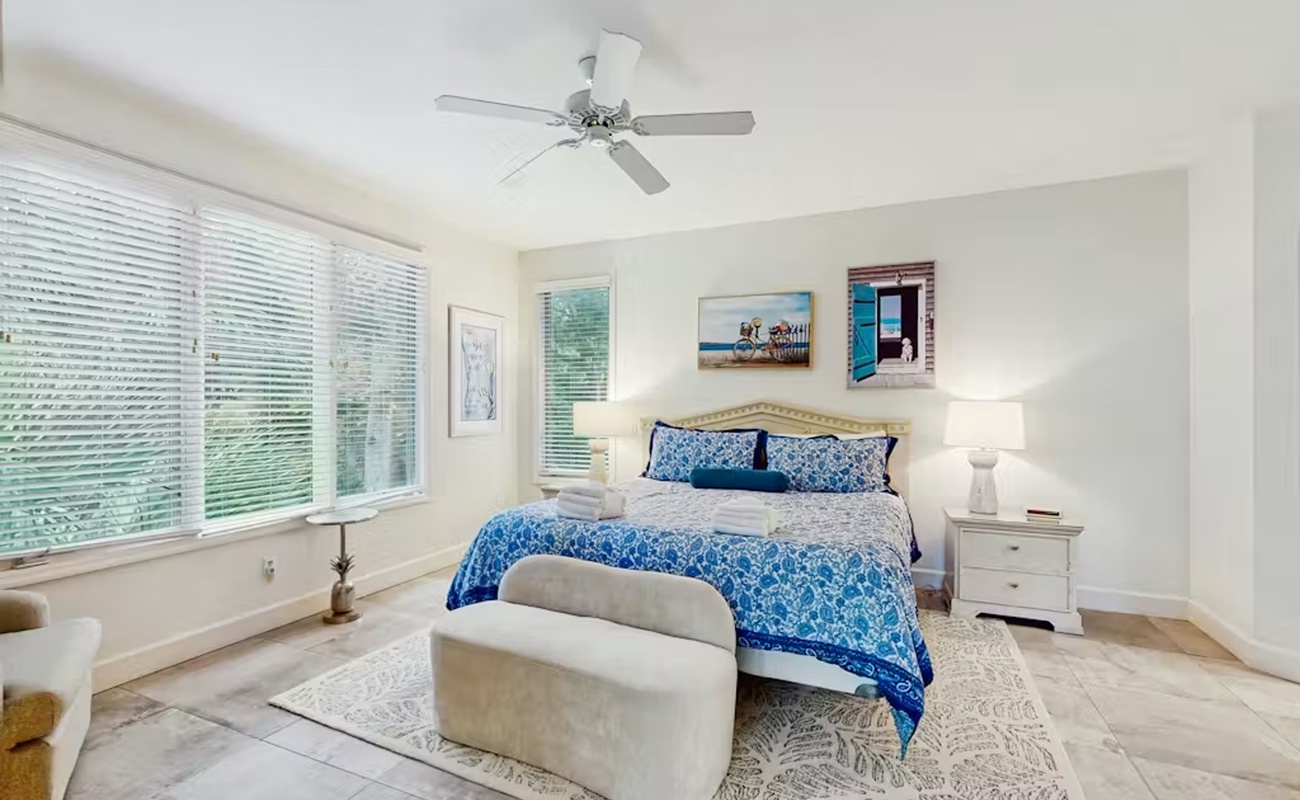 Unwind in your serene bedroom featuring ocean-inspired blue linens, gentle ceiling fan breezes, and peaceful garden views through large windows.