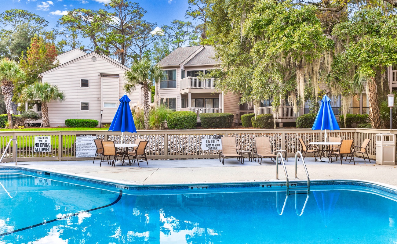 Sparkling community pool with lounge chairs and umbrellas, surrounded by lush tropical landscaping and residential buildings.