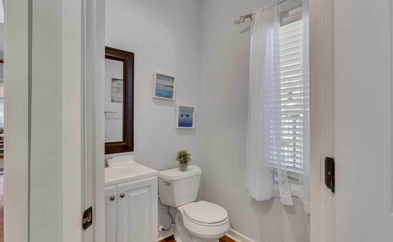 Your private powder room features crisp white finishes and coastal artwork, creating a peaceful retreat with natural light streaming through shuttered windows.
