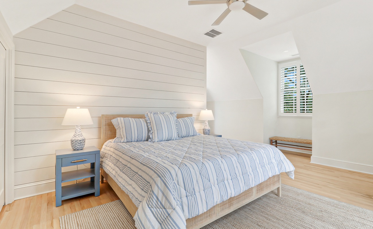 Drift off to sleep in your bright, coastal-inspired bedroom with soft striped linens and warm natural light filtering through shuttered windows.