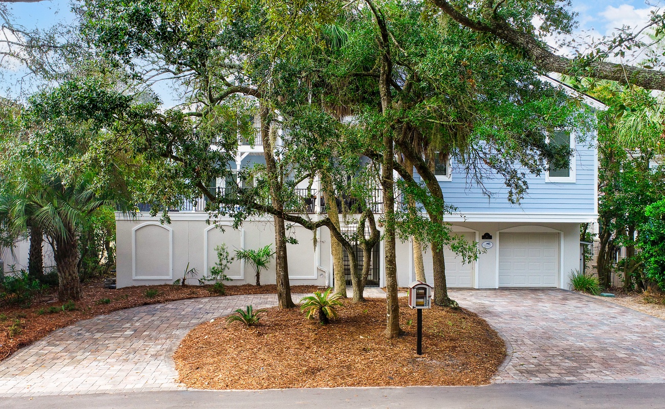 Modern white vacation home nestled among mature oak trees with circular brick driveway and tropical landscaping.