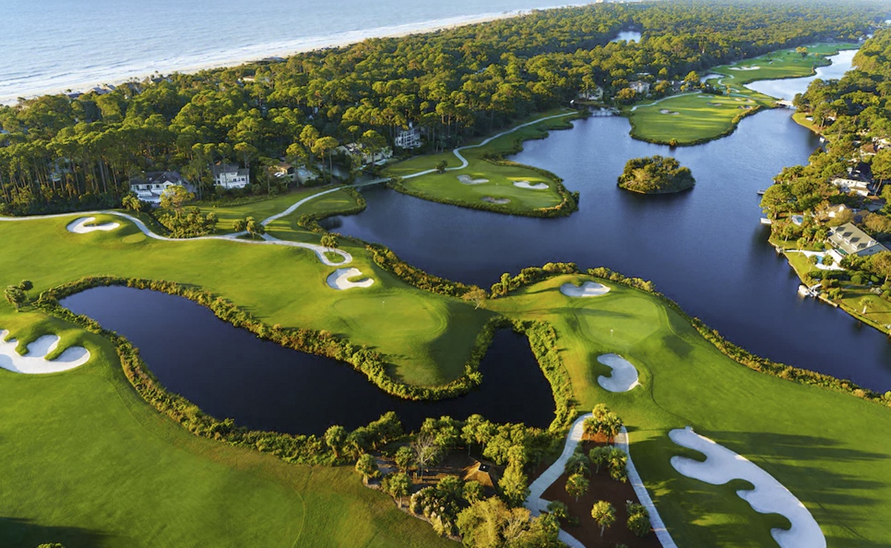Aerial view of championship golf course with water features winding through pristine greens near the coastline.