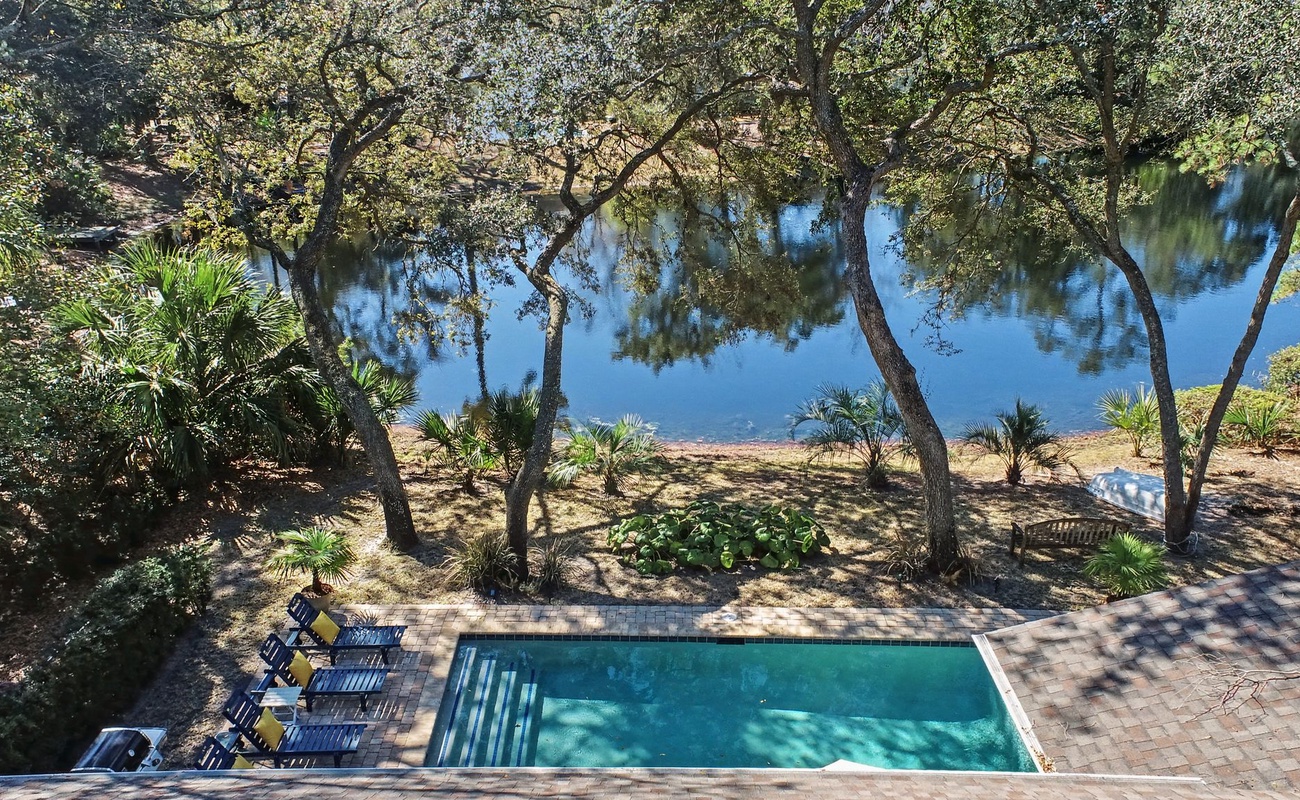 Aerial view of the property's sparkling pool area surrounded by mature trees and landscaped gardens, with waterfront access nearby.