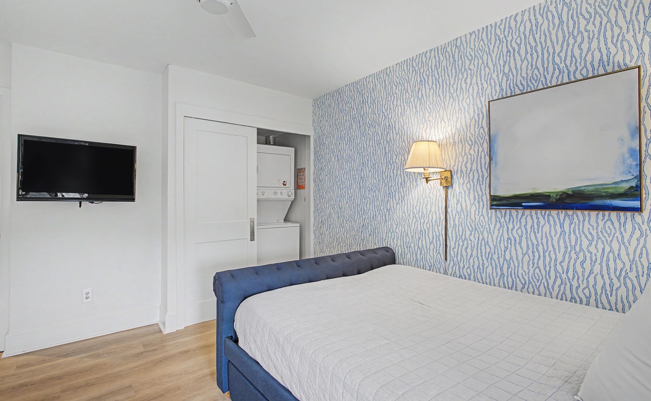 Relax in your serene bedroom with coastal-inspired decor, featuring a comfortable bed, wall-mounted TV, and convenient in-room laundry facilities.