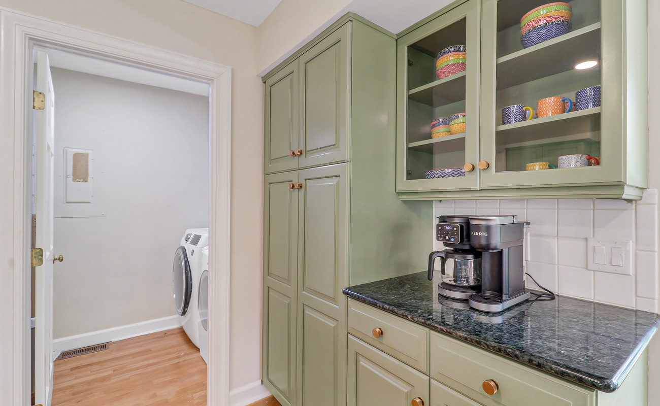 Your charming kitchenette features sage green cabinetry and granite counters, perfect for brewing morning coffee with colorful dishes displayed in glass-front cabinets.