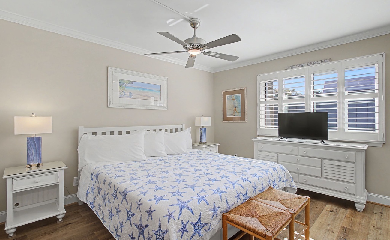 Relax in your coastal-inspired bedroom with soft starfish bedding, gentle ceiling fan, and natural light streaming through plantation shutters.
