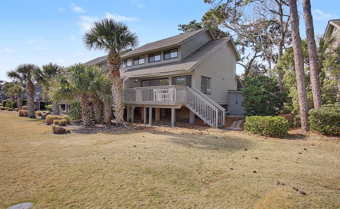 A charming coastal home nestled among tall pines and palm trees, featuring a spacious deck and natural landscaping in a peaceful residential setting.