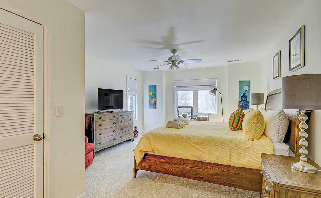 Relax in your bright, cheerful bedroom with golden linens and coastal touches. The ceiling fan keeps you cool while colorful artwork adds personality.