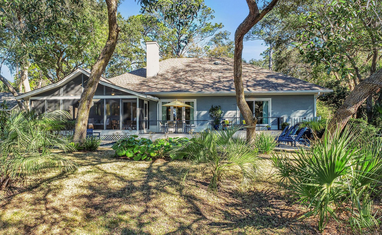 A charming single-story home nestled among mature trees and tropical landscaping, featuring a welcoming covered patio perfect for outdoor relaxation.