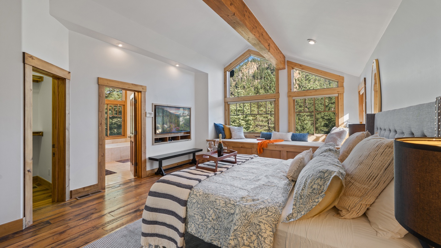 Wake to natural light streaming through tall windows into your spacious master suite, where rustic wood beams and cozy seating create the perfect mountain retreat for relaxation.