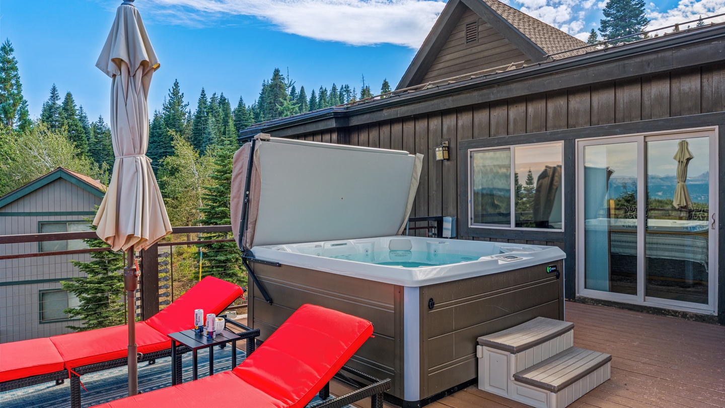 Soak in your private hot tub on the deck, surrounded by towering pines and blue skies. Two lounge chairs and a shaded umbrella invite you to relax between dips, with the peaceful mountain setting just beyond.