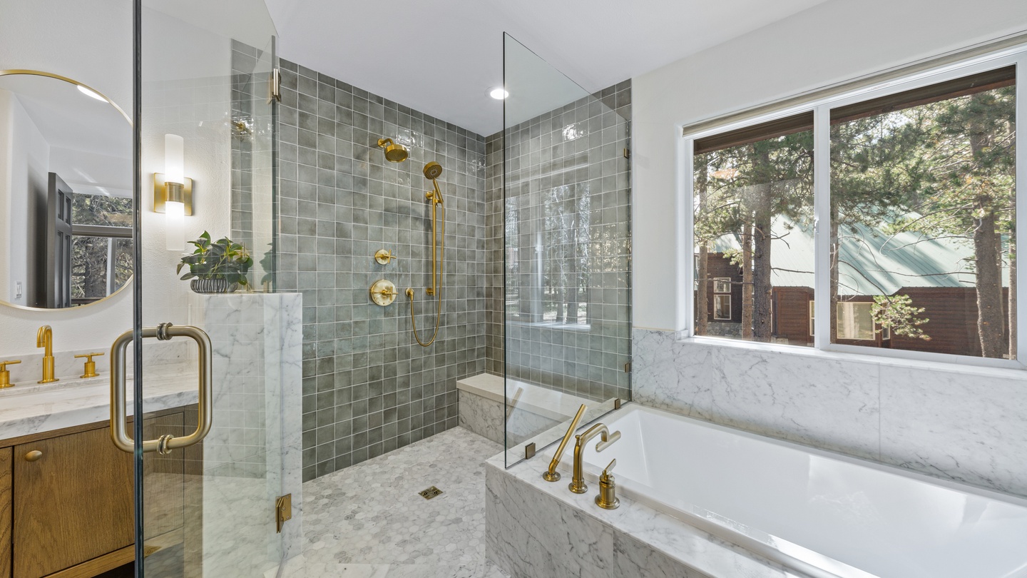 Luxurious marble and sage tile create a spa-like retreat with a soaking tub, glass-enclosed shower with gold fixtures, and natural light streaming through tall windows overlooking the pines.