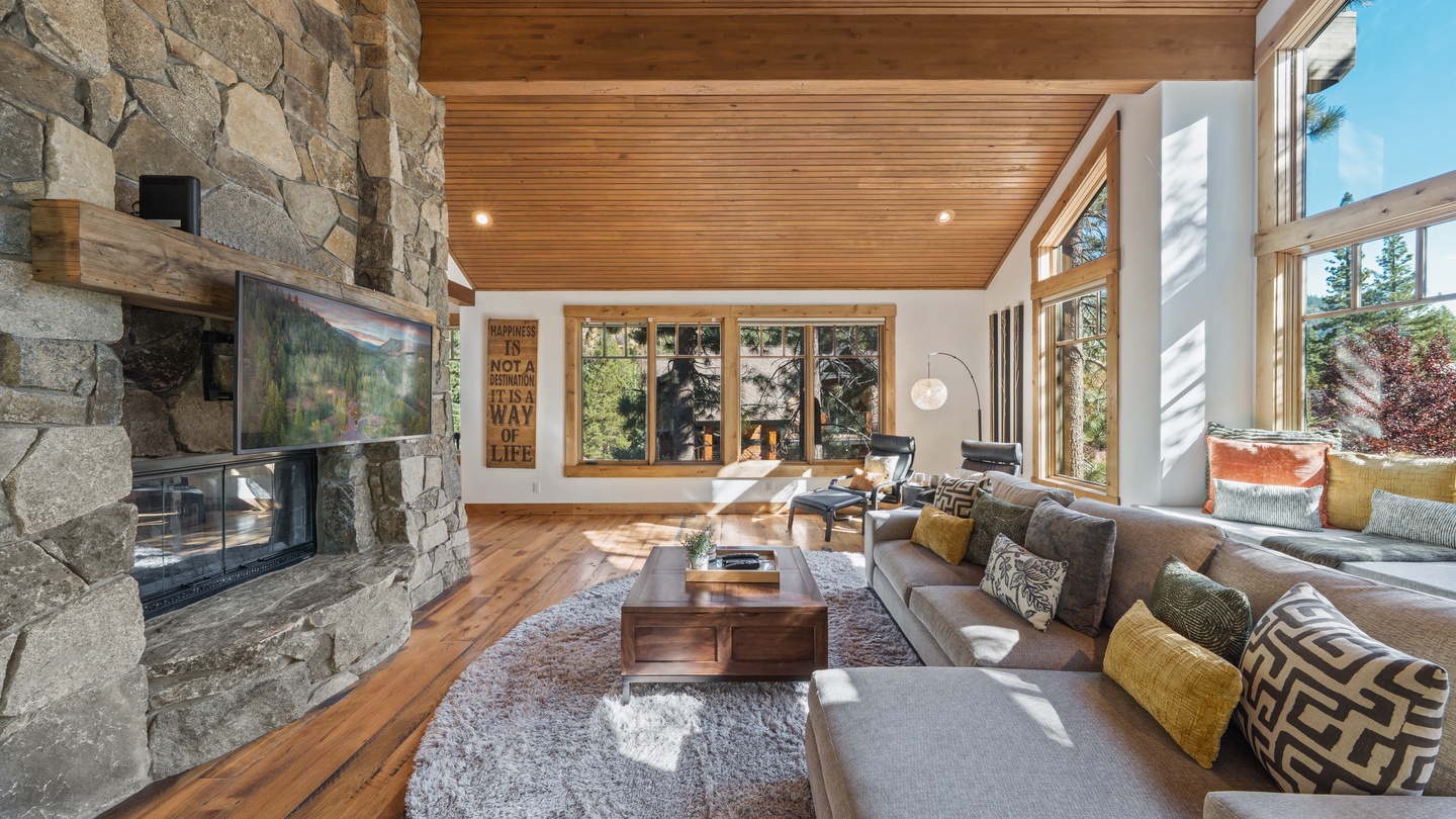 Sink into plush seating beneath soaring wood-beam ceilings, where sunlight streams through floor-to-ceiling windows framing forest views. Your retreat centers on a stunning stone fireplace, perfect for cozy mountain evenings.