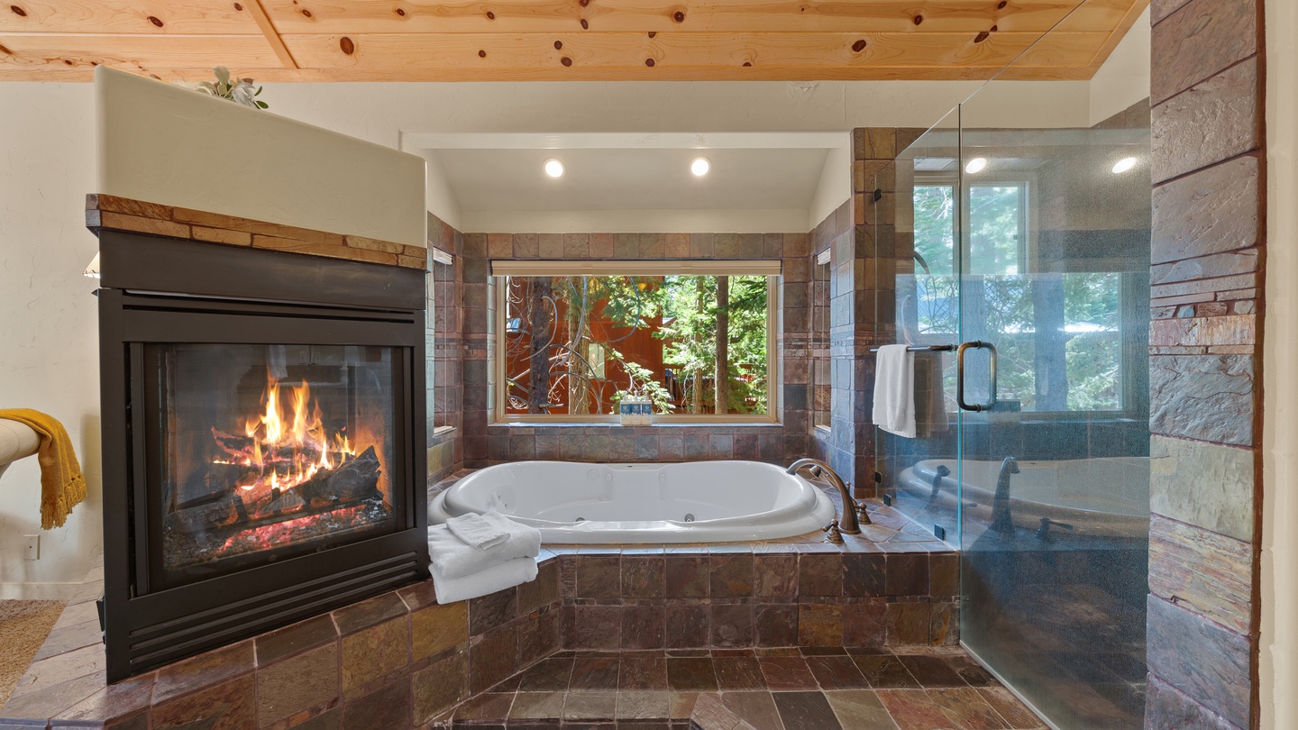 Unwind in your luxurious bathroom with a soaking tub beside a cozy fireplace and a spacious walk-in shower, all framed by natural forest views through the window.