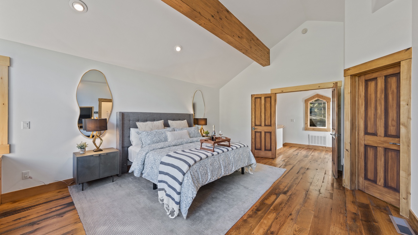 Unwind in your serene bedroom featuring vaulted ceilings with rustic exposed beams, plush bedding, and warm wood floors that create a cozy mountain retreat atmosphere.