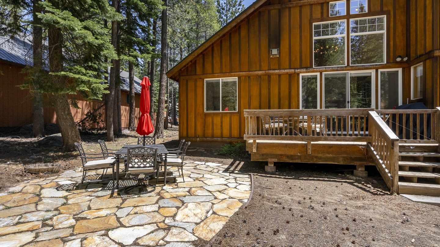 A warm wood cabin nestled among towering pines features a spacious deck and stone patio with outdoor dining, perfect for mountain mornings and evenings under the trees.