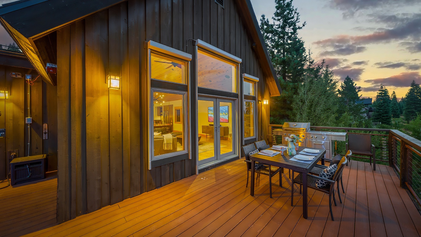 A modern cabin exterior with deck dining space set against a dramatic sunset sky, surrounded by towering evergreens and natural forest landscape.