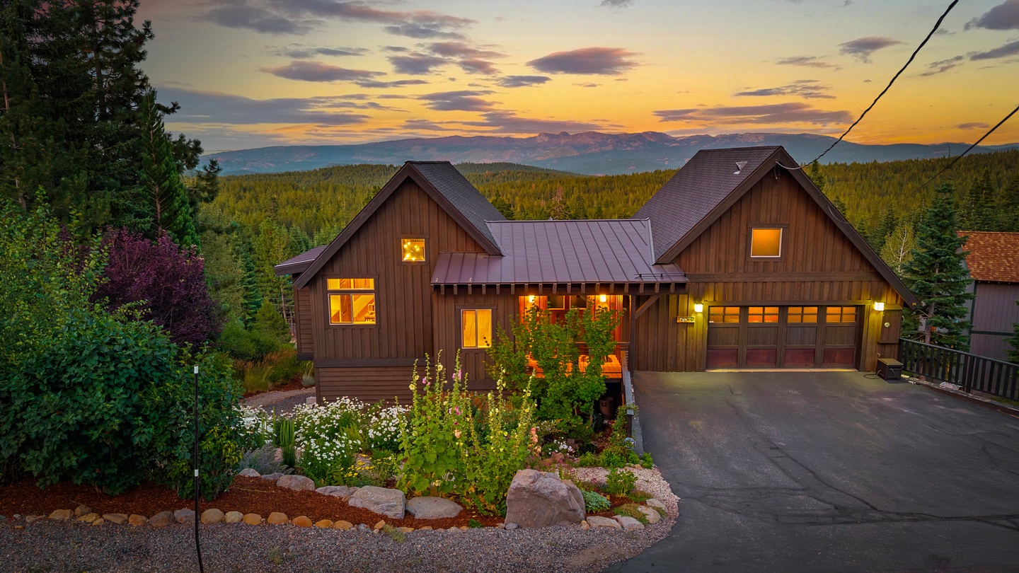 This inviting mountain retreat sits nestled among towering pines with stunning sunset views over the surrounding forest. The warmly lit exterior welcomes guests to a peaceful escape surrounded by natural beauty and seasonal landscaping.
