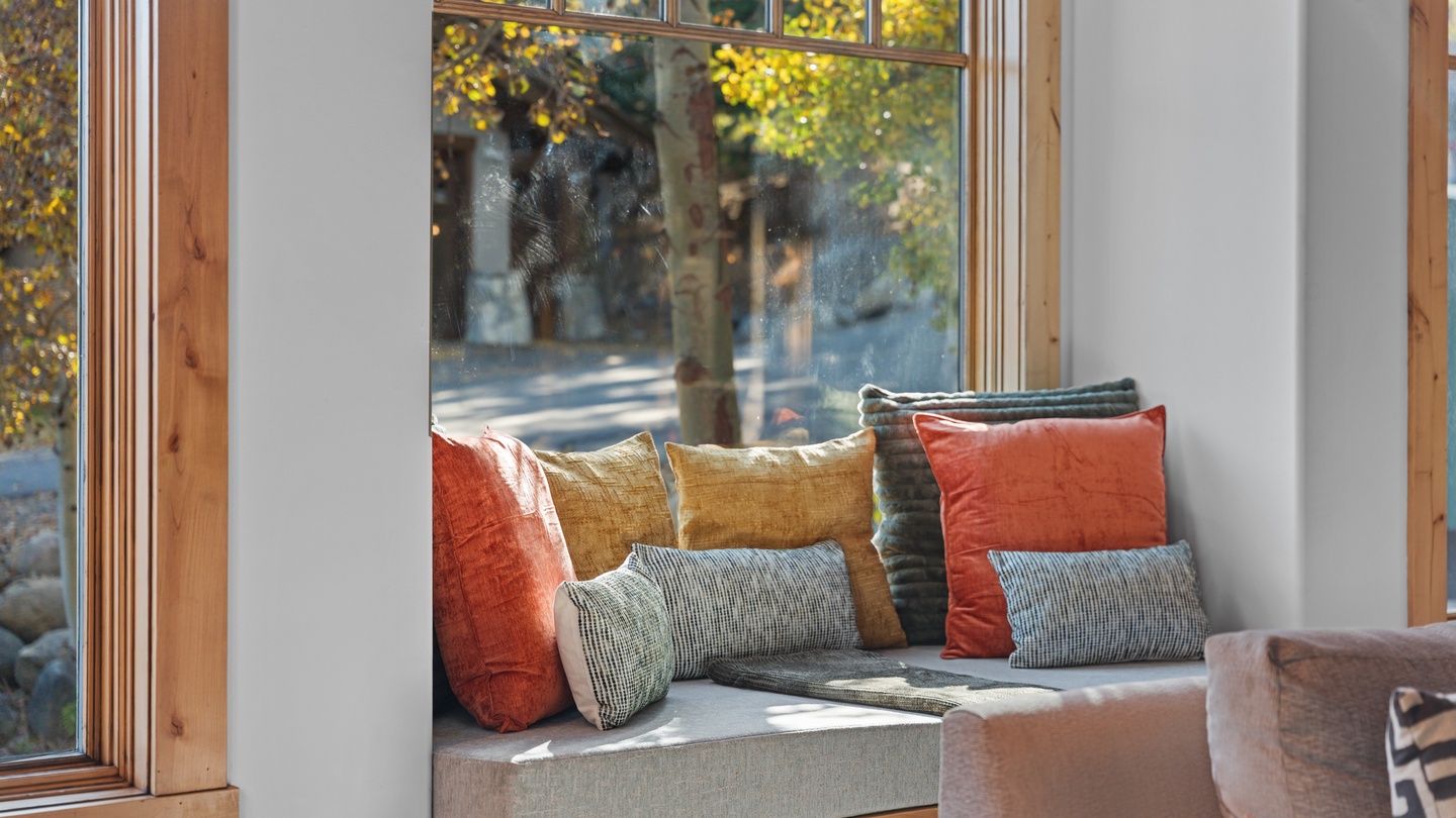 Cozy up in your window seat with colorful cushions and soak in the sunlight streaming through the tall windows, showcasing vibrant autumn foliage just beyond the glass.