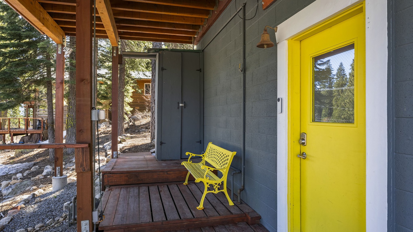 Charming covered entrance with cheerful yellow door and seating area surrounded by towering pines.