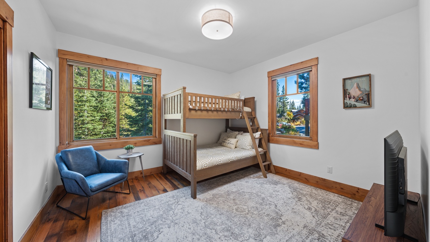 Sunlight streams through generous windows framing forest views in your cozy bedroom retreat. Warm wood tones and a comfortable bunk bed create the perfect space for restful mountain getaways.