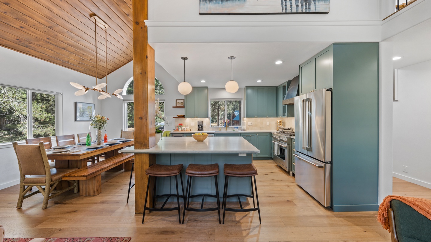 Bright, open-concept kitchen and dining space with sleek teal cabinetry and warm wood tones. Natural light streams through large windows, creating the perfect welcoming atmosphere for cooking and gathering.