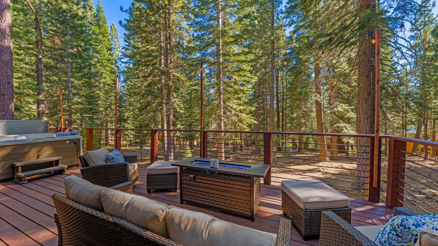 The surrounding forest is visible from this spacious outdoor deck. The area features comfortable seating and a grill for outdoor dining beneath towering pines and blue sky.