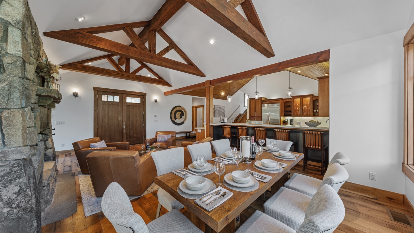 Soaring timber beams frame an open-concept great room with a farmhouse dining table, plush leather seating, and a stone fireplace bathed in natural light from craftsman-style doors.