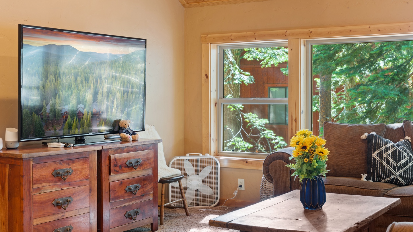 Unwind in this warm living room with wood-paneled ceilings, comfortable seating, and natural light streaming through windows overlooking the trees. Your perfect space to relax after a day exploring.