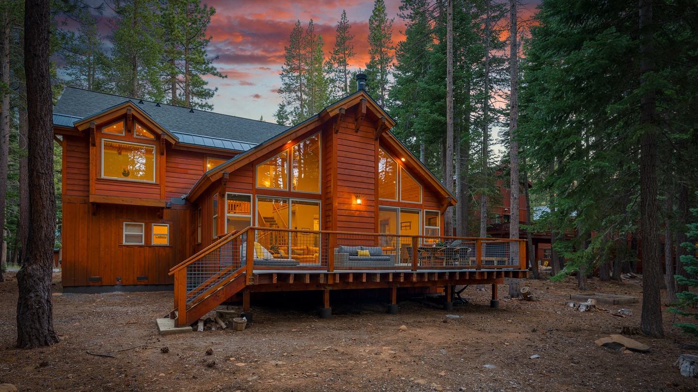 A stunning mountain retreat nestled among towering pines captures a vibrant sunset through floor-to-ceiling windows, while the warm glow from within highlights the inviting living space just beyond the spacious deck.