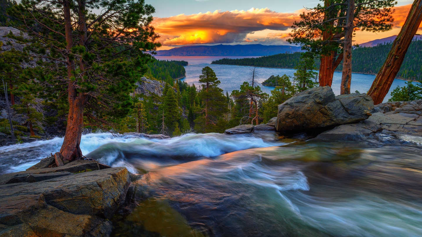 A stunning natural landscape near the property features a rushing mountain river framed by towering pines, with a dramatic sunset illuminating the lake and forested peaks beyond.