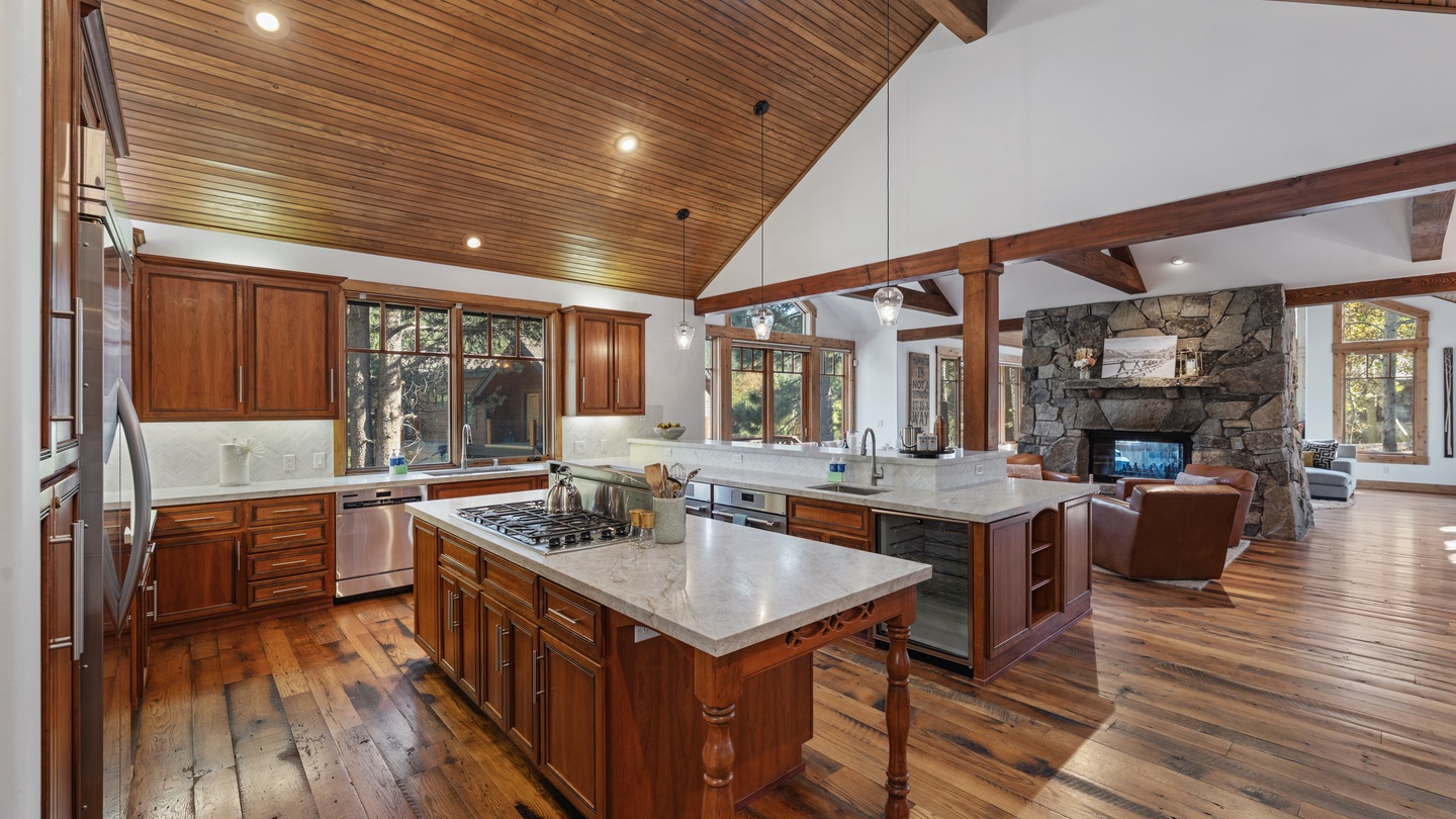 Rich wood tones and open-beam ceilings bring warmth to this inviting kitchen and living space, where you can cook, relax by the stone fireplace, and soak in natural light streaming through expansive windows.
