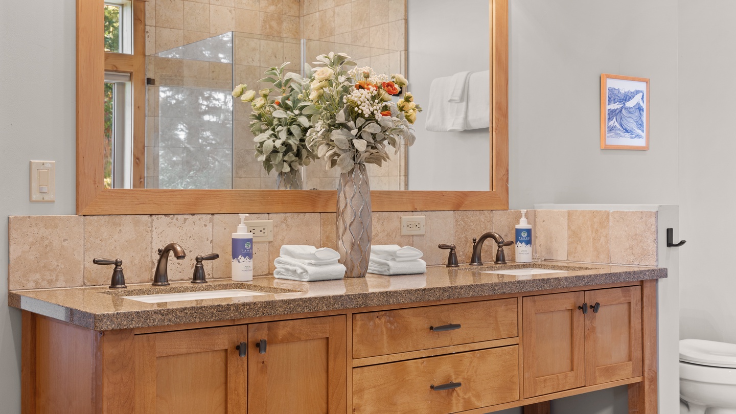 This beautifully appointed bathroom offers two sinks with warm wood cabinetry, soft natural lighting from the window, and fresh linens ready for your stay.