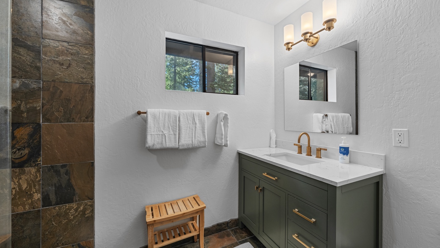 Refresh in your stylish bathroom with warm brass fixtures, a spacious walk-in shower with textured stone tile, and forest views through the window.