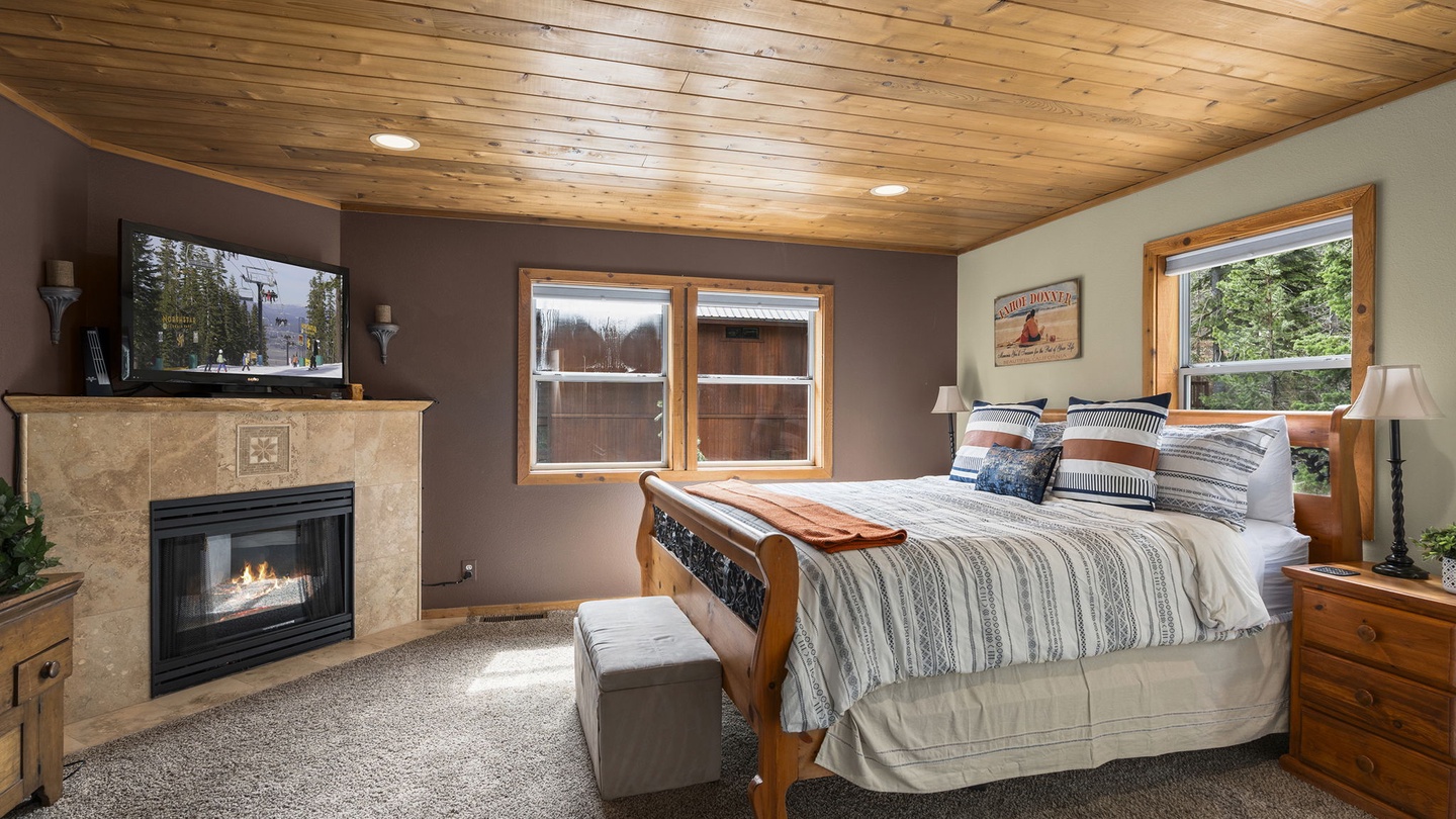 Cozy up in this warm mountain bedroom featuring a crackling fireplace, rustic wood ceiling, and peaceful forest views.